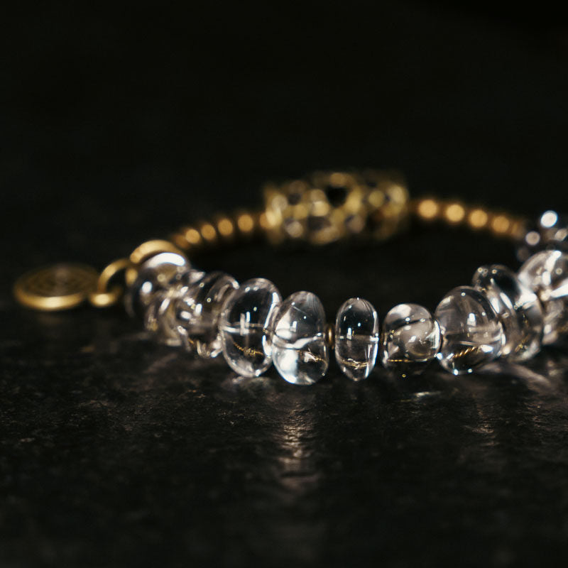 Macro photography of a clear beaded bracelet showing light-reflecting crystal beads and a detailed vintage-style patterned brass focal bead.