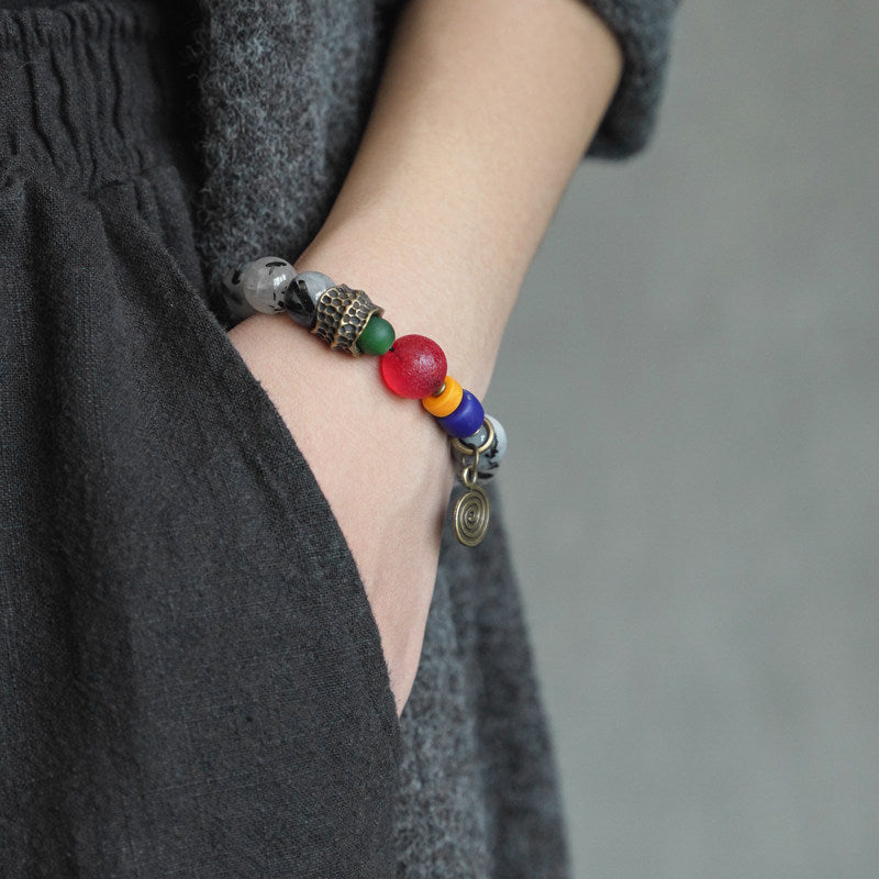 A person wearing a bohemian ethnic-style beaded bracelet with a vintage brass spiral charm, styled with a grey textured linen outfit.