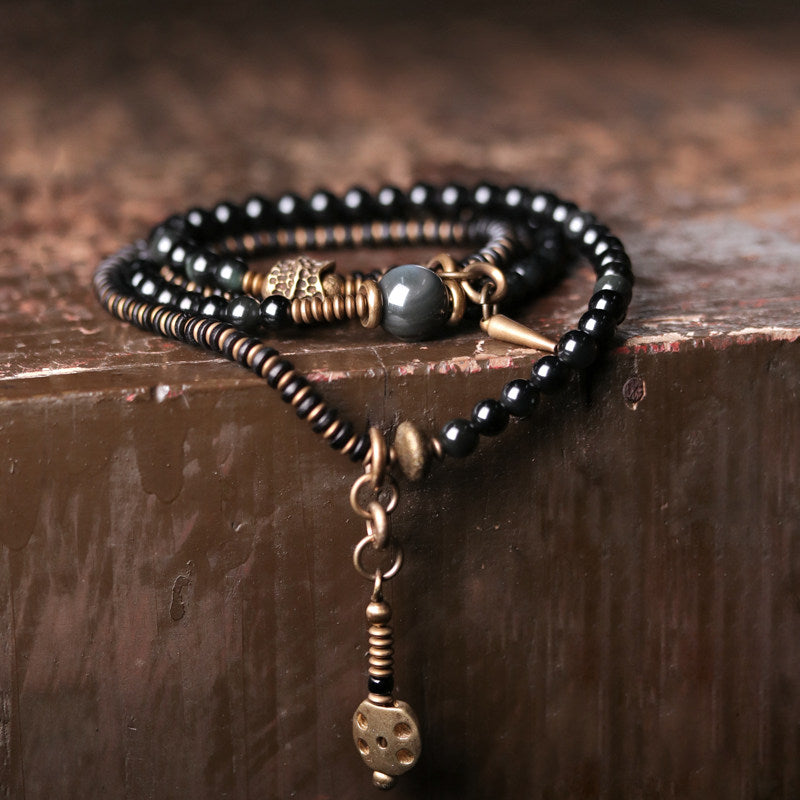 A multi-strand wrap bracelet featuring glossy black beads, antique brass hardware, and a spike pendant, draped over a rustic dark wood surface.