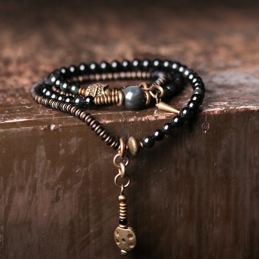 A multi-strand wrap bracelet featuring glossy black beads, antique brass hardware, and a spike pendant, draped over a rustic dark wood surface.