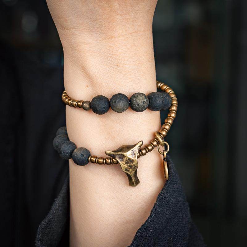 A person wearing a Southwestern-style bracelet stack combining matte black volcanic stones and distressed brass beads with a bull horn pendant.