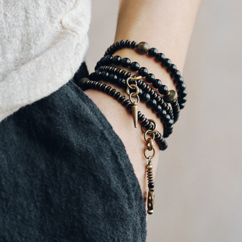A person wearing a layered black onyx beaded bracelet stack with a vintage brass spike pendant, paired with a dark textured linen outfit.