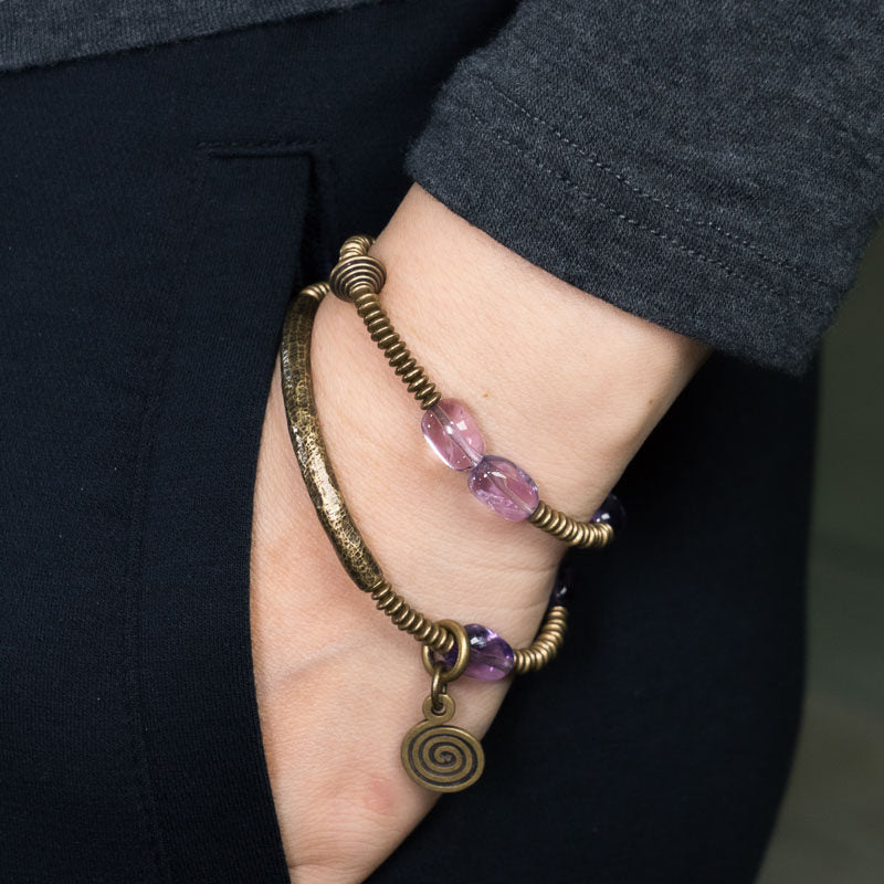 Lifestyle shot of a person wearing the Serene Lucky Royal Wisdom bracelet, showcasing the stacked look of purple amethyst beads and sculptural brass tubes against a minimalist background.