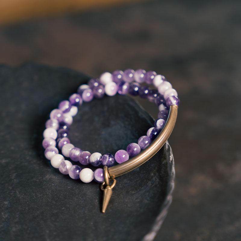 Serene Lucky brand banner featuring an amethyst multi-wrap bracelet with textured brass tubes and a spiral sun talisman, emphasizing spiritual healing and artisanal quality.