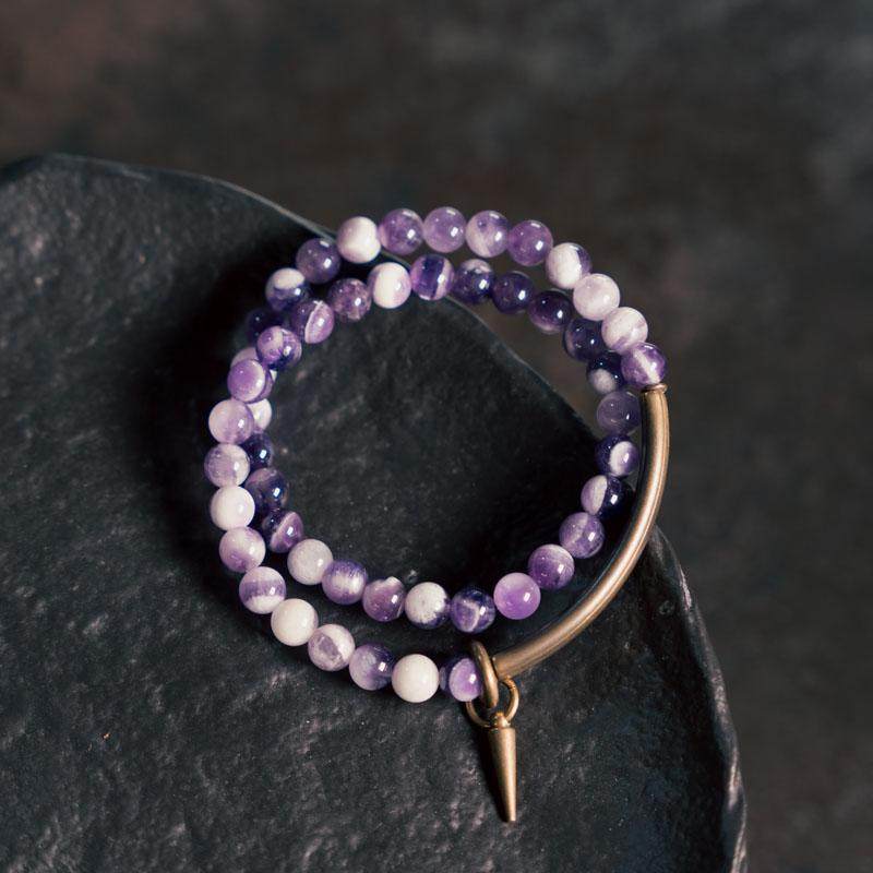 Side view of Serene Lucky wrap bracelet, showcasing tiered layers of polished purple amethyst beads and elongated antique brass tubes with a soft bokeh background.