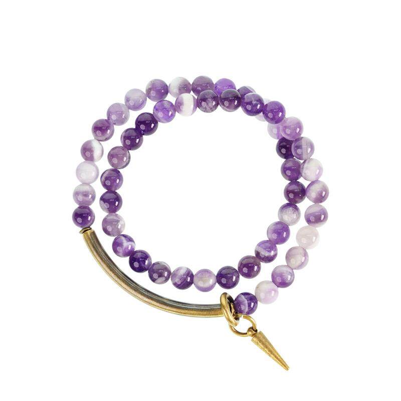 A high-angle flatlay of the Serene Lucky multi-wrap bracelet, showcasing deep purple amethyst beads, curved brass tubes, and a spiral sun charm on a textured dark background.