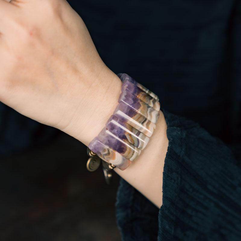 Macro detail of the Serene Lucky bracelet showcasing a hand-polished amethyst slab link and the intricate texture of the antiqued brass spiral sun talisman.