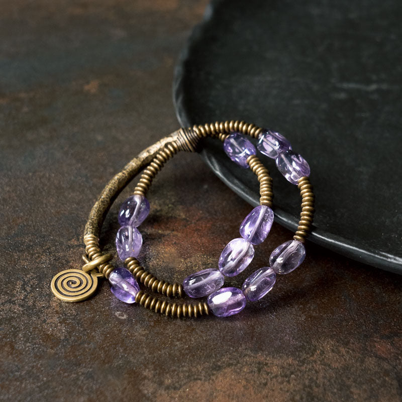 Top view of Serene Lucky Royal Wisdom wrap bracelet, featuring four layers of purple amethyst beads and an antiqued brass coin charm on a dark textured background.