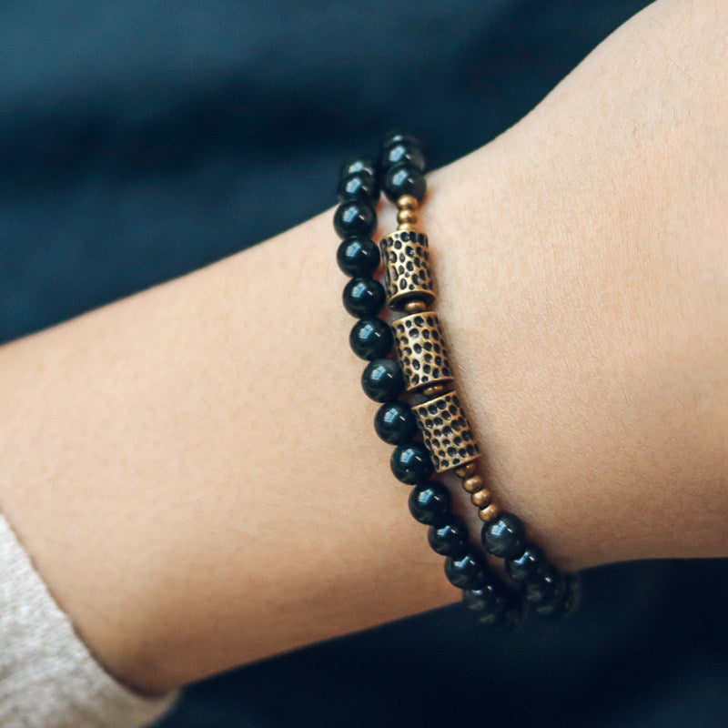 Lifestyle shot of Serene Lucky's Celestial Weaver bracelet on a woman's wrist, featuring hollow lattice brass spheres and polished black obsidian beads.