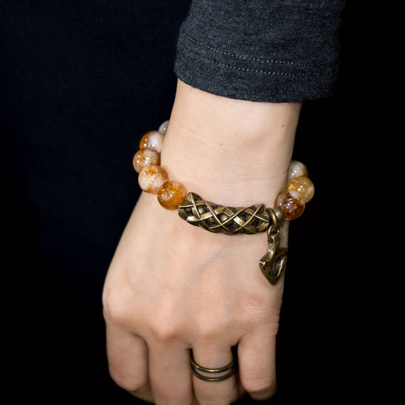 A side-view lifestyle shot of the Serene Lucky solar talisman bracelet, featuring a prominent woven-texture brass cylinder and glowing yellow citrine beads on a wrist with a black sleeve.