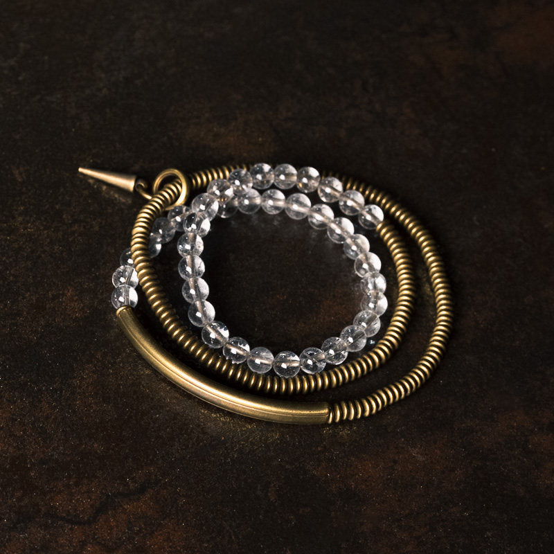 A macro close-up of Serene Lucky multi-wrap bracelet, showing the contrast between smooth brass tubes, coiled metal textures, and translucent clear quartz beads on a dark background.