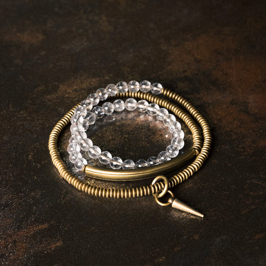 An aesthetic low-light shot of the Serene Lucky multi-wrap bracelet, featuring luminous clear quartz beads and antiqued brass tubes on a dark textured surface.