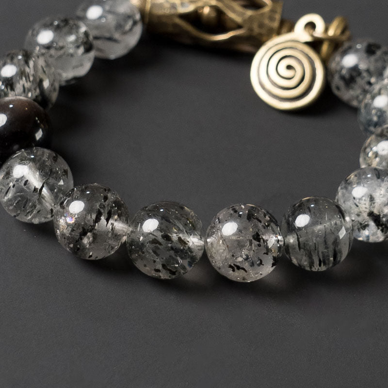 Detailed macro shot of the Serene Lucky Cosmic Shield bracelet, highlighting the intricate floral-etched brass cylinders and the iconic spiral charm on a textured background.