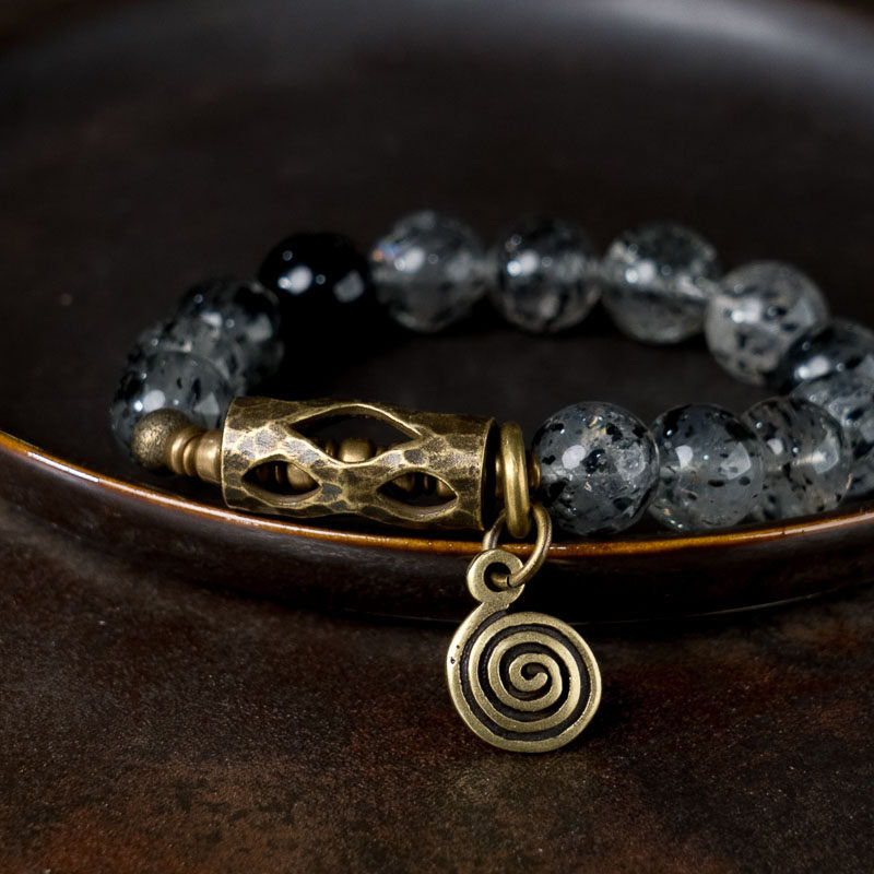 Macro view of Serene Lucky Cosmic Shield bracelet, focusing on the intricate floral engravings on antiqued brass cylinders and the central spiral energy symbol.
