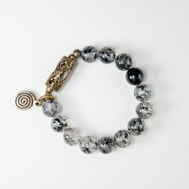 A person wearing the Serene Lucky Cosmic Shield bracelet, showcasing the reflective silver sheen obsidian beads and detailed brass floral accents against a dark knit sleeve.