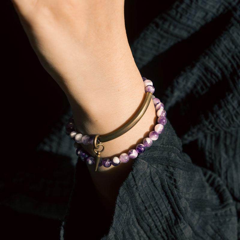 Close-up of Serene Lucky multi-wrap bracelet showing vibrant purple amethyst beads and intricate textured brass tube accents against a moody, dark background.