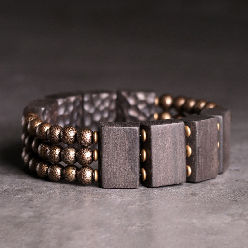 Close-up of Serene Lucky architectural bracelet featuring hand-carved Ebony wood slabs and textured antiqued brass beads on a dark background.