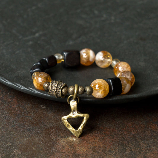 An artistic shot of the Serene Lucky Golden Clarity bracelet, featuring faceted yellow citrine beads, dark ebony wood cubes, and a handcrafted hammered brass triangle charm on a slate background.