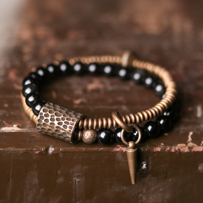 Artistic close-up of Serene Lucky's Midnight Fortress bracelet, focusing on the honeycomb-textured brass link and glossy obsidian beads in a moody, low-light setting.