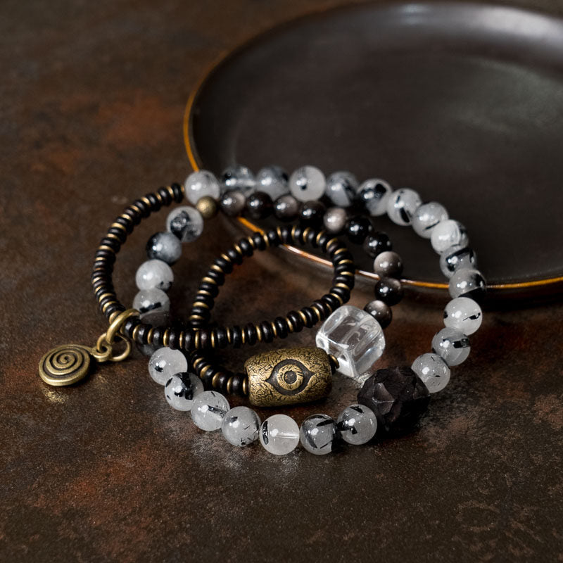 A handcrafted Serene Lucky multi-layer wrap bracelet featuring dark Ebony wood, shimmering Silver Sheen Obsidian, and Clear Quartz with architectural antiqued brass accents on a minimalist background.