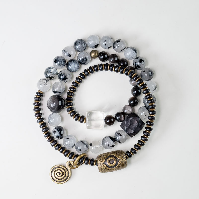 A Serene Lucky multi-layer wrap bracelet featuring dark Ebony wood, shimmering Silver Sheen Obsidian, Black Rutilated Quartz, and Clear Quartz beads with antiqued brass structural accents.