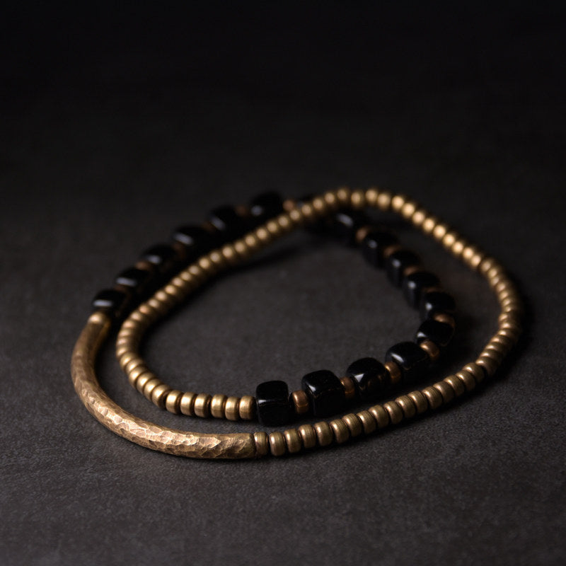 A rustic flat-lay of the Serene Lucky Night Rhythm bracelet on dark wood, featuring square obsidian beads and brass spikes alongside raw stones.