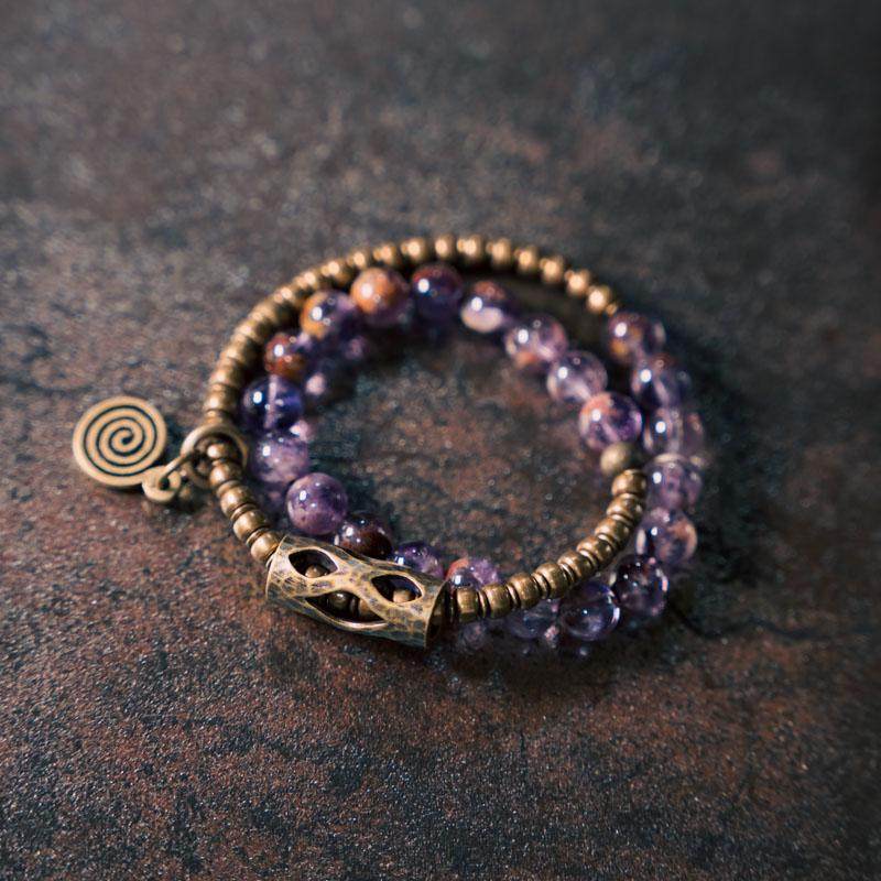 Serene Lucky brand poster featuring a purple phantom quartz multi-wrap bracelet with textured brass tubes and a spiral sun charm on a black background.
