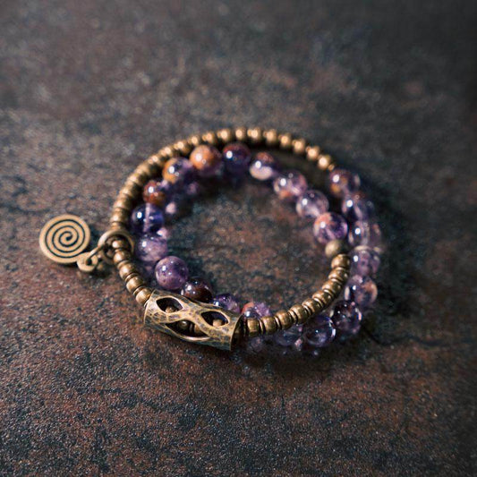 Serene Lucky brand poster featuring a purple phantom quartz multi-wrap bracelet with textured brass tubes and a spiral sun charm on a black background.