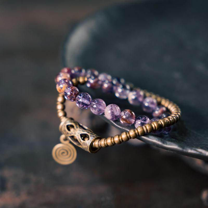 A high-angle flatlay of the Serene Lucky multi-wrap bracelet, showcasing layers of purple phantom quartz beads and cylindrical matte brass tubes on a neutral grey background.