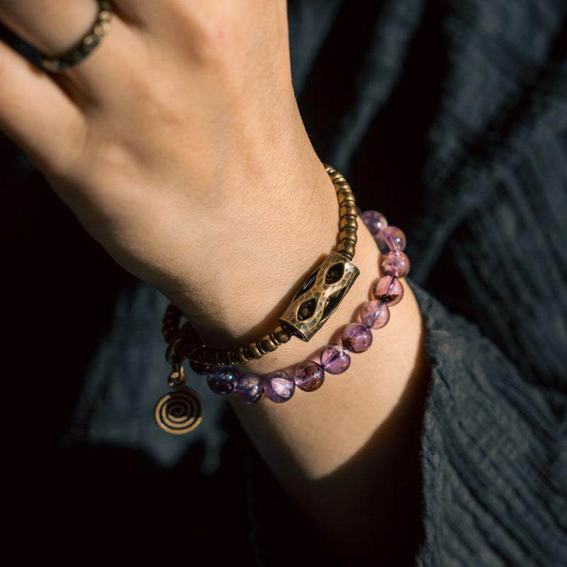 A side-angle macro shot of the Serene Lucky wrap bracelet, showcasing the unique phantom inclusions in purple quartz beads and the textured antique brass tubes.