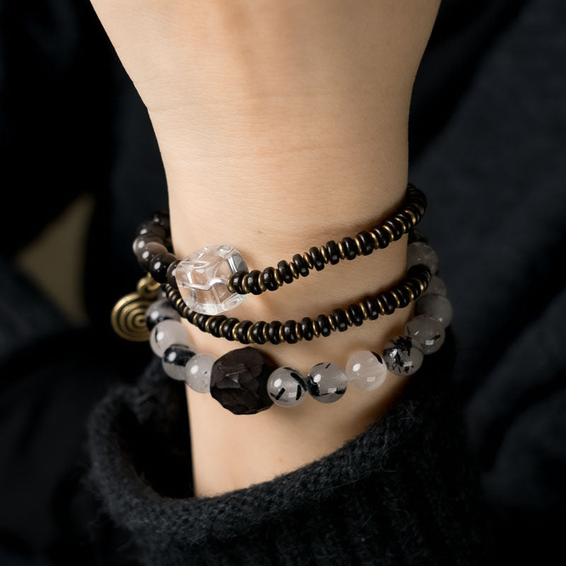 Macro shot of the Serene Lucky Shadow & Light bracelet, highlighting the contrast between dark Ebony wood, needle-like Black Rutilated Quartz, and shimmering Silver Sheen Obsidian with antiqued brass spring coils.