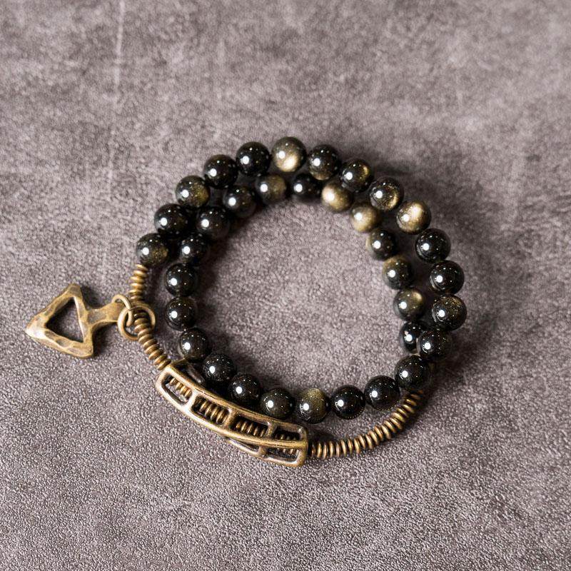 A cinematic shot of Serene Lucky's Solar Gears bracelet on a grey slate surface, highlighting the shimmering golden sheen obsidian and brass gear accents.
