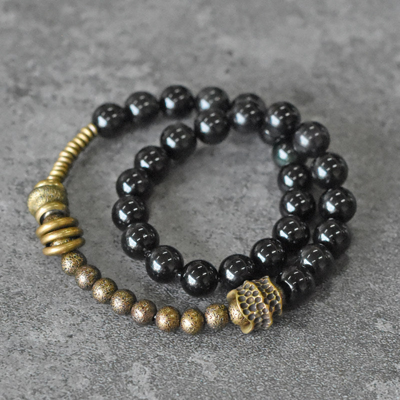 A lifestyle shot of Serene Lucky's Tectonic Force bracelet featuring molten-textured brass accents and black obsidian beads on a rustic wood background.