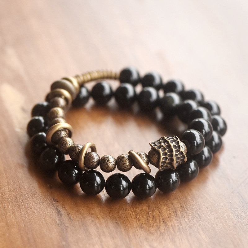 High-angle shot of Serene Lucky's Tectonic Force bracelet resting on a rustic wood surface, showing the contrast of molten-textured brass and black obsidian beads.