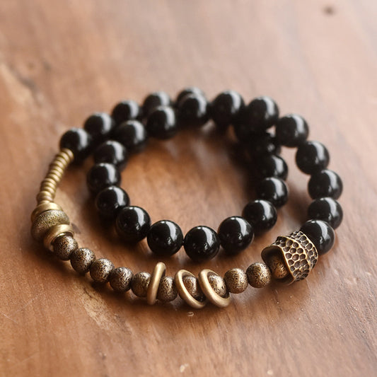 A professional flat-lay of Serene Lucky's Tectonic Force bracelet on a warm wood surface, highlighting the molten-textured brass and black obsidian beads.