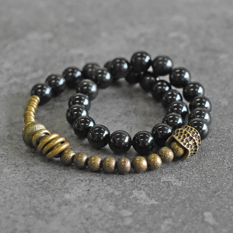 A professional flatlay of Serene Lucky's Tectonic Force wrap bracelet, showcasing molten-textured brass accents and glossy obsidian beads on a dark wood surface.