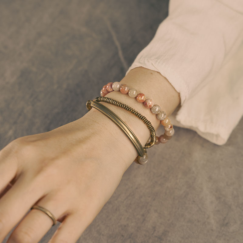 A multi-layer Serene Lucky bracelet called Timeless Echo, featuring orange Fossil Coral beads, curved brass tubes, and a spiral pendant worn on a wrist.