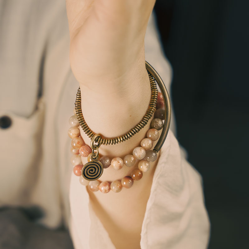 A multi-layer Serene Lucky bracelet called Timeless Echo, featuring orange Fossil Coral beads, curved brass tubes, and a spiral pendant worn on a wrist.