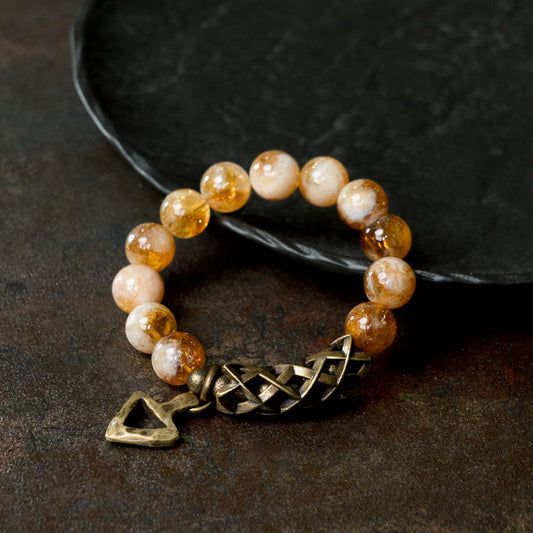 A close-up of a person wearing the Serene Lucky Solar Path bracelet, highlighting the handcrafted brass triangle charm and woven metal tube against faceted citrine beads.