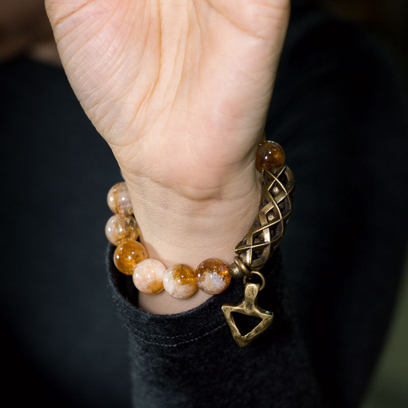 Close-up of the Serene Lucky Solar Path bracelet on a wrist, featuring a brass triangle charm hanging from a woven-patterned metal tube with faceted yellow citrine beads.