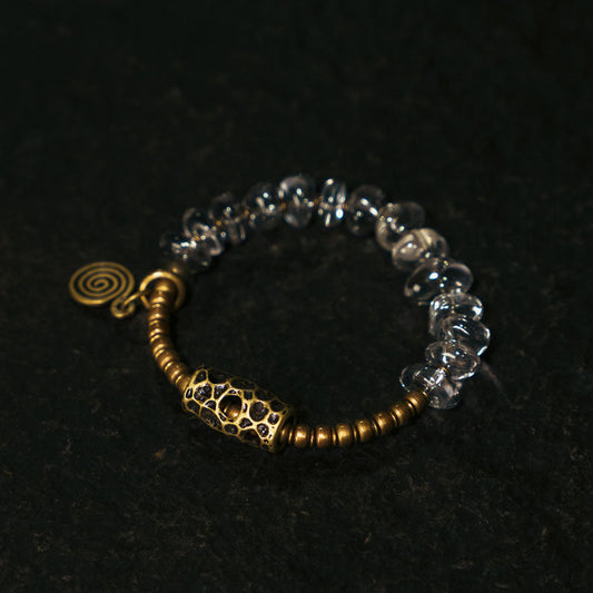 Detailed view of a handmade beaded bracelet with clear crystal rondelle beads and antique-style hammered brass accents, including a spiral charm, lying on a black textured background.