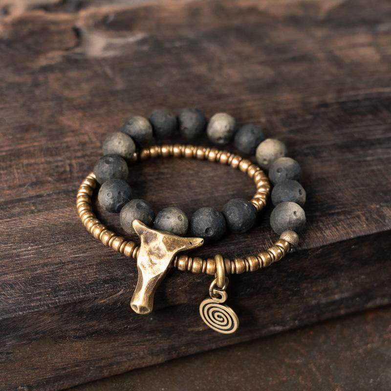 A rustic jewelry stack featuring a black volcanic stone beaded bracelet and a hammered antique brass bead chain with a bull head charm.