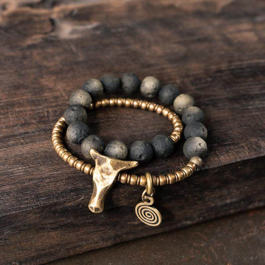 A rustic jewelry stack featuring a black volcanic stone beaded bracelet and a hammered antique brass bead chain with a bull head charm.